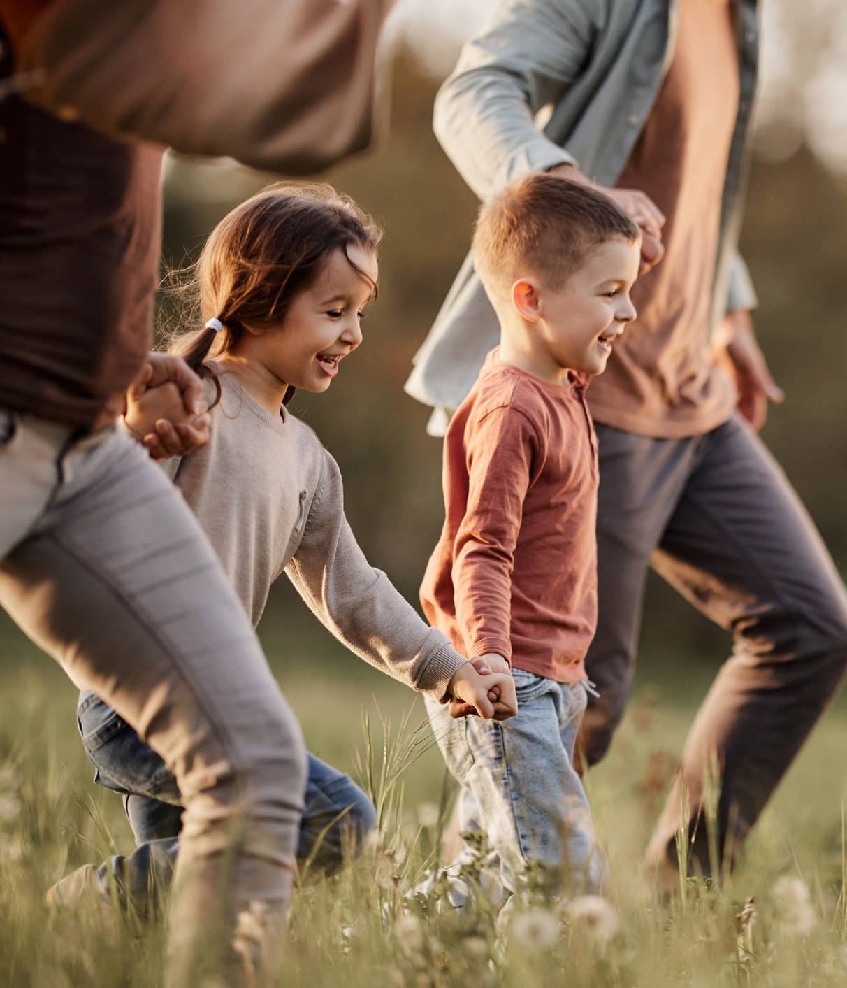 Two siblings, a boy and a girl, about 5 years old, hold hands with each other and their parents while running through a field. They are thriving due to south island family support.